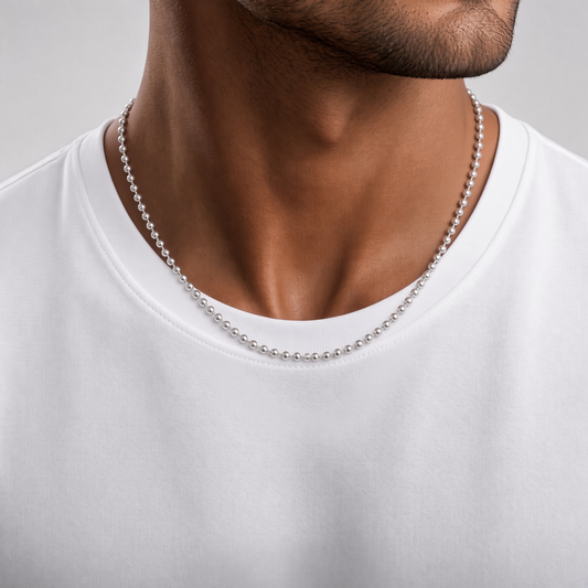 A close-up of a man's neck and upper chest wearing a silver ball chain necklace over a white t-shirt, set against a neutral gray background.